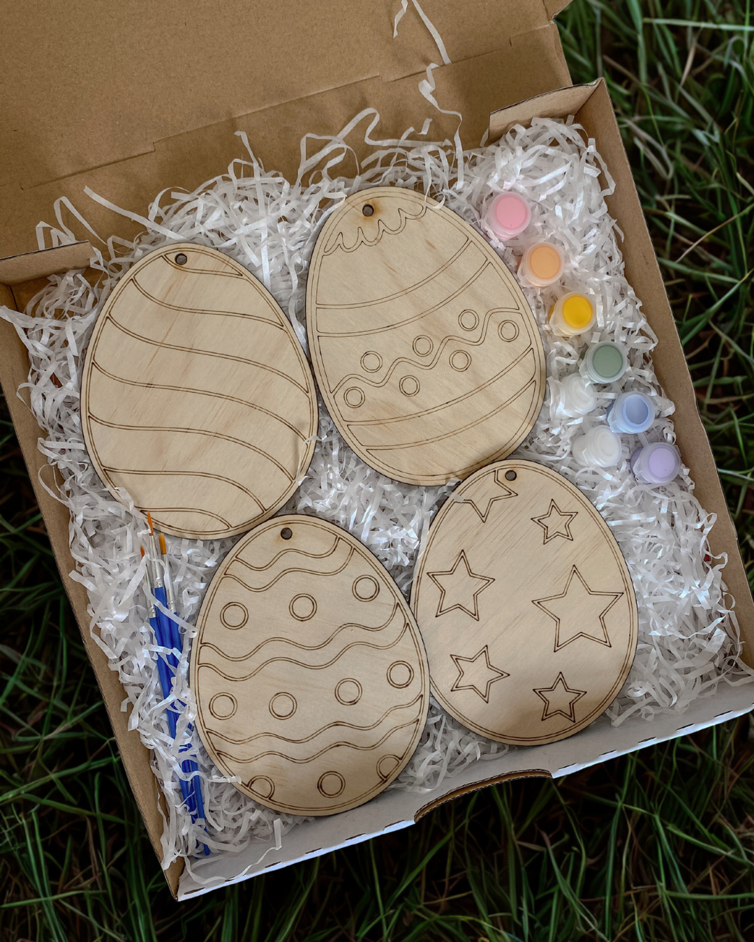 Wooden Easter egg ornaments with star and circle patterns in a box with craft supplies.