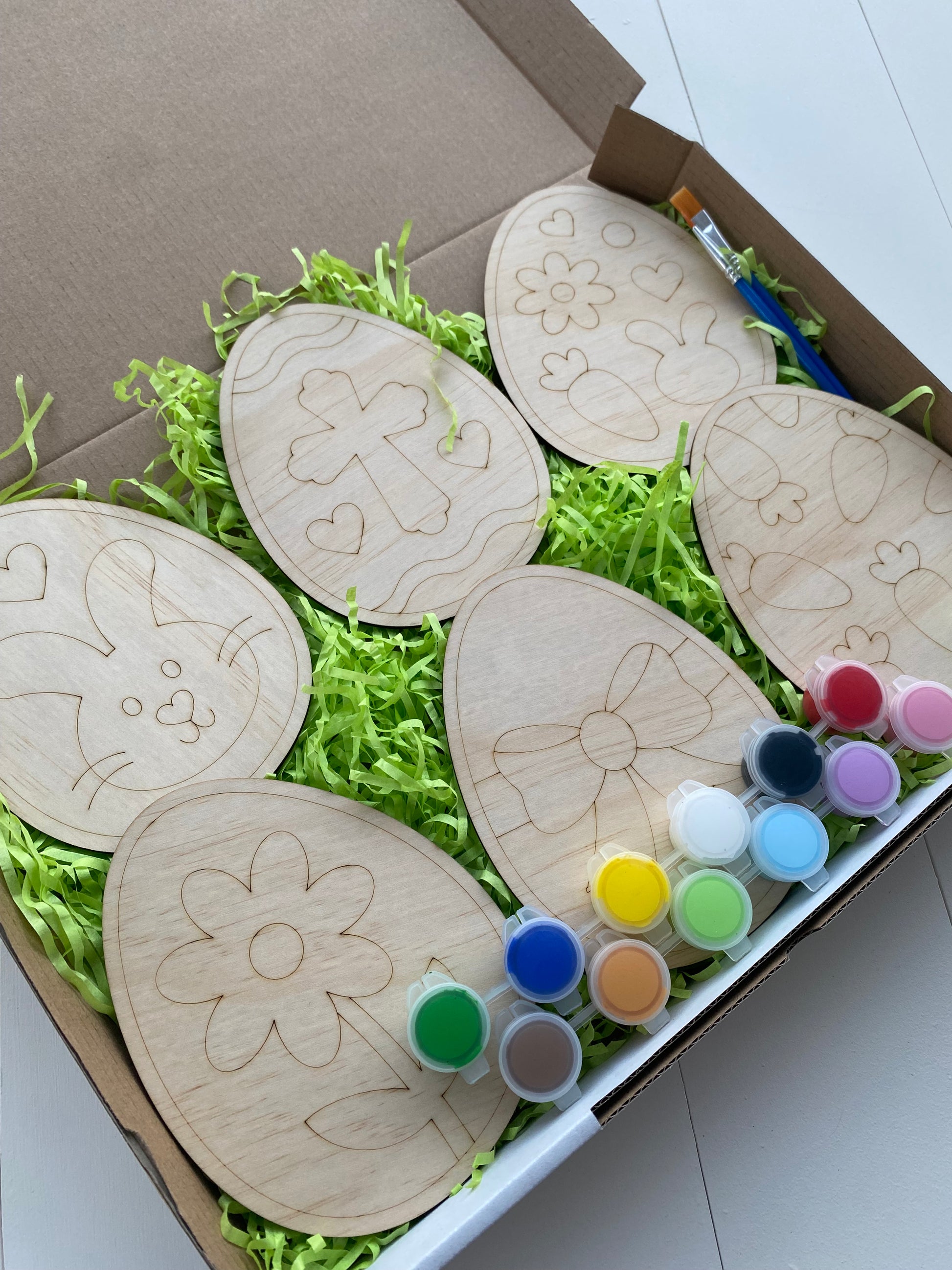 Wooden Easter egg-shaped crafts with paint bottles on a green shredded paper background